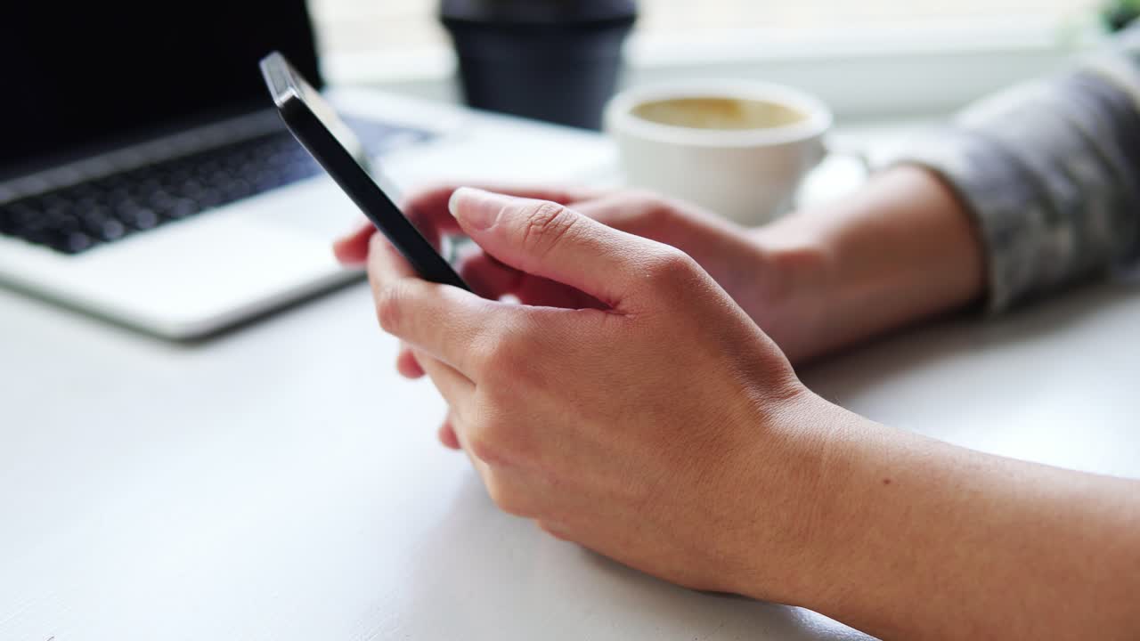 Close Up view of female hands using app on smartphone while sitting in cafe with a cup of coffee and laptop on the table