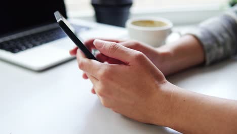 Vista-De-Cerca-De-Las-Manos-Femeninas-Usando-La-Aplicación-En-El-Teléfono-Inteligente-Mientras-Se-Sienta-En-La-Cafetería-Con-Una-Taza-De-Café-Y-Una-Computadora-Portátil-Sobre-La-Mesa