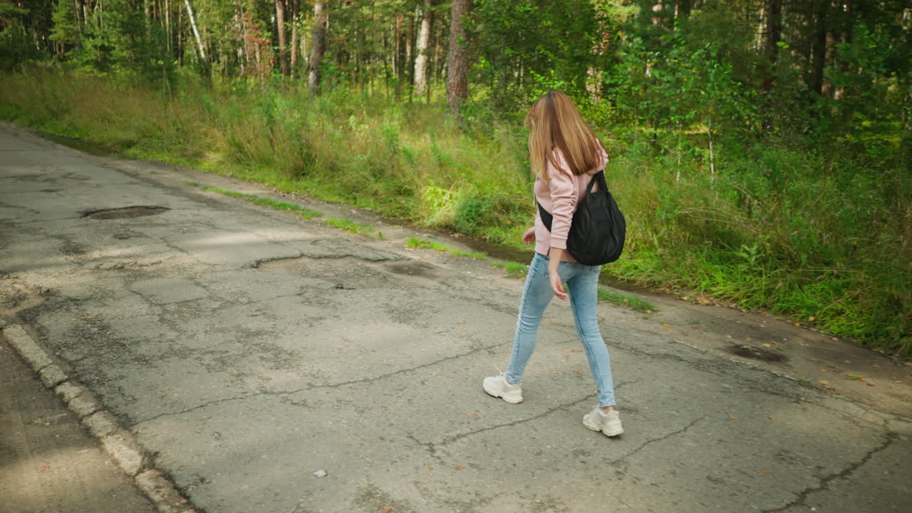 Young lady with loose hair walking thoughtfully along forest road carrying black backpack, surrounded by lush greenery and tall trees, calm mood with sunlight casting soft shadows on cracked pavement