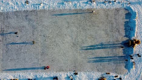 winter aerial birds view hold over multi use manmade ice rink at snow covered park sunset as parent teaches child to skate with triangle support others practice hockey another trys figure skating 2-3