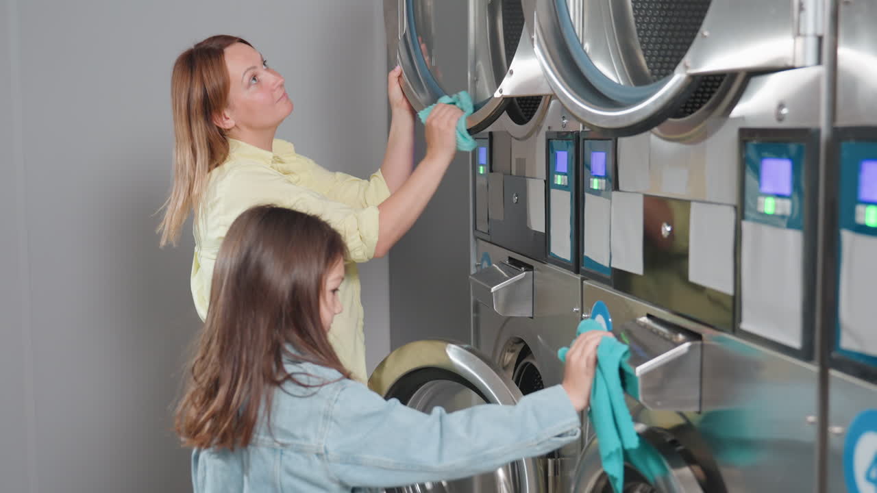School girl in denim jacket wipes dryer door with turquoise cloth beside mother inside laundromat, assisting cleanup across stainless machines, focus on hygiene, routine maintenance, calm teamwork