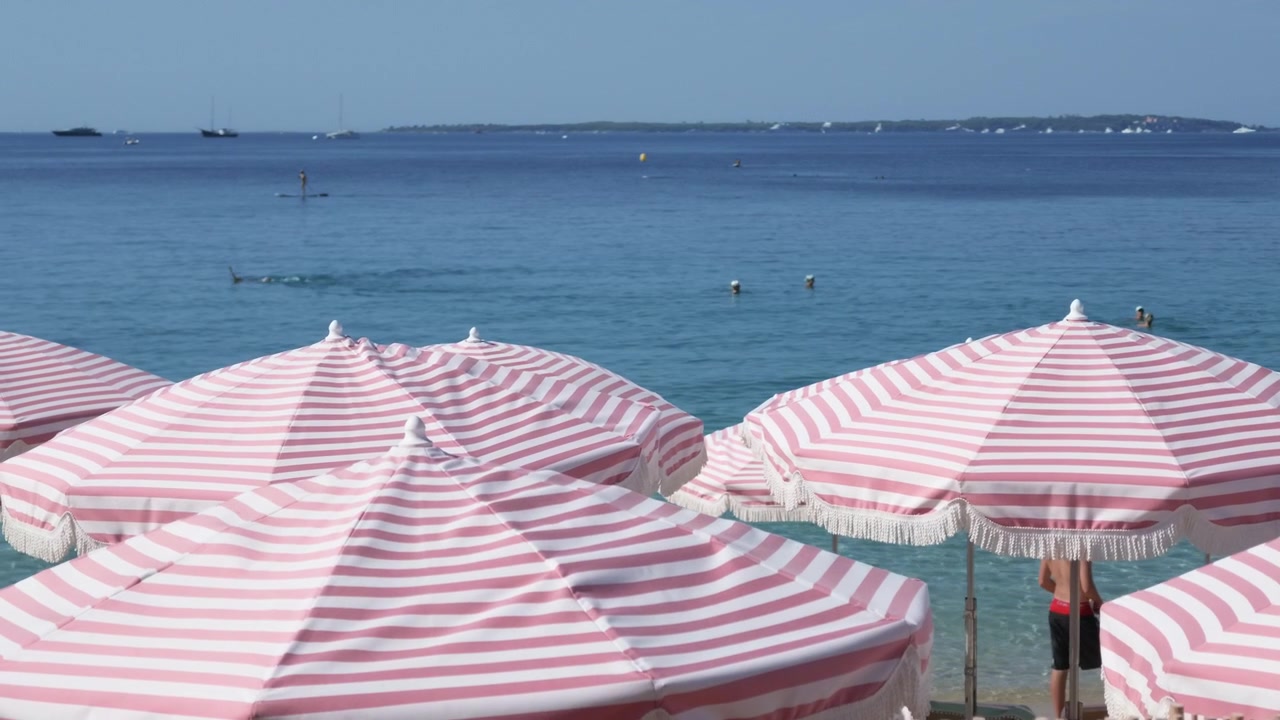 Primo piano di ombrelloni a strisce rosa e bianche con bagnanti e un paddleboarder in mare, Cannes, Francia