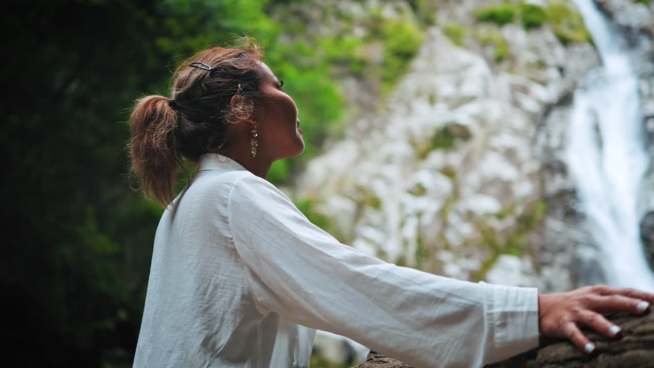 joven modelo femenina asiática mira a la cascada mientras se apoya en una barandilla de madera