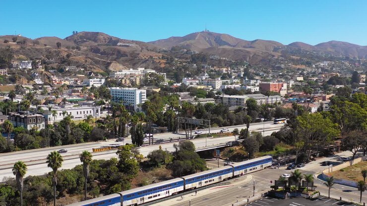 Una ripresa aerea con drone del treno Amtrak Pacific Surfline che passa attraverso la città costiera della California meridionale, Ventura, California, con l'autostrada in primo piano e le montagne sullo sfondo.
