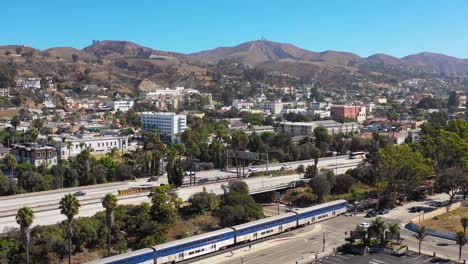 a drone aerial of the pacific surfline amtrak train passing through the southern california beach town of ventura california with freeway foreground and mountains background