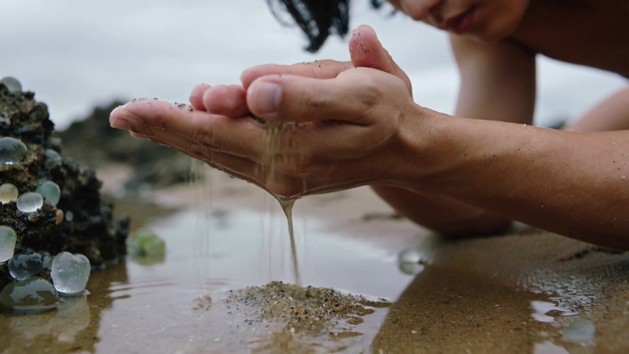 Crouching shirtless man cupping wet sand, inspecting draining water forming mound near gel beads