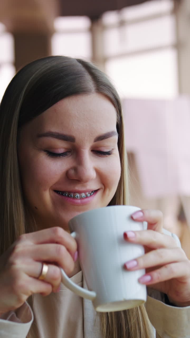 Happy smiling lady enjoys rest and tea during lunch break. Young woman looks dreamily into the window. Vertical video