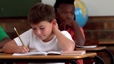 Little-boy-writing-in-notepad-in-classroom-and-smiling-at-camera