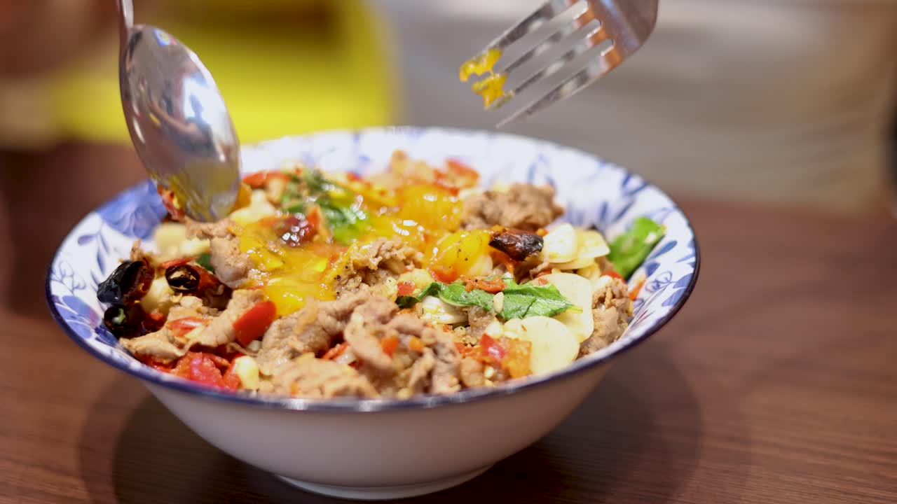 A person uses a spoon and fork to break and mix a runny egg over a colorful Thai rice dish in bright indoor lighting