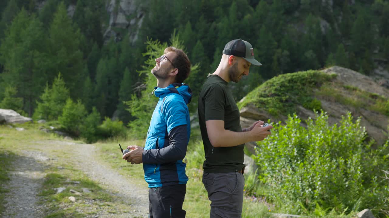 Due piloti di droni in piedi schiena contro schiena in un suggestivo scenario montano, concentrati sui loro droni. Il paesaggio verde lussureggiante e l'ambiente naturale creano una scena ideale per un'avventura all'aperto