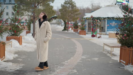 girl in warm beige winter coat with black hoodie and earmuffs looks up at sky while calmly turning behind, snowy path lined with festive trees and decorations creates a serene