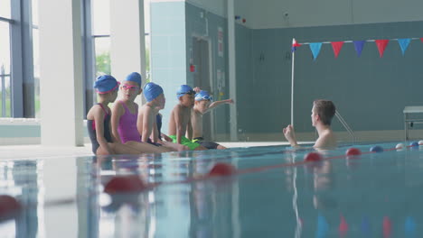 male coach in water gives children instructions in swimming class as they sit on edge of indoor pool