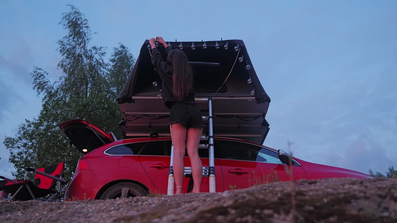 Woman prepares campsite with glowing lights on red car at peaceful twilight