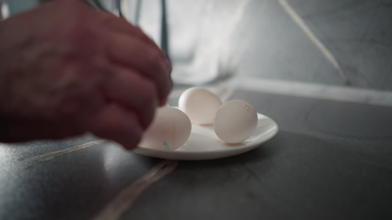 Elderly woman in green sweatshirt placing egg onto plate in kitchen. Close-up shot capturing hands and focused action in modern kitchen, reflecting daily routine and self-sufficiency