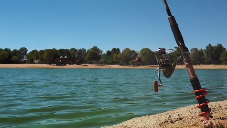 fishing rod stuck in sandy shore of rippling lake on sunny day