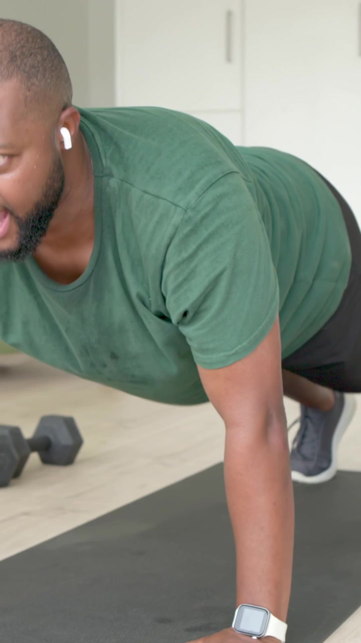 Vertical video: Starting set, man in green tee doing push-ups on home mat with dumbbells ending set