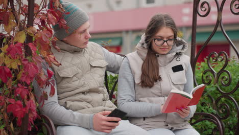 young couple sitting closely on park bench in autumn, man holding phone while woman reads book, surrounded by red leaves, dressed warmly in casual winter wear