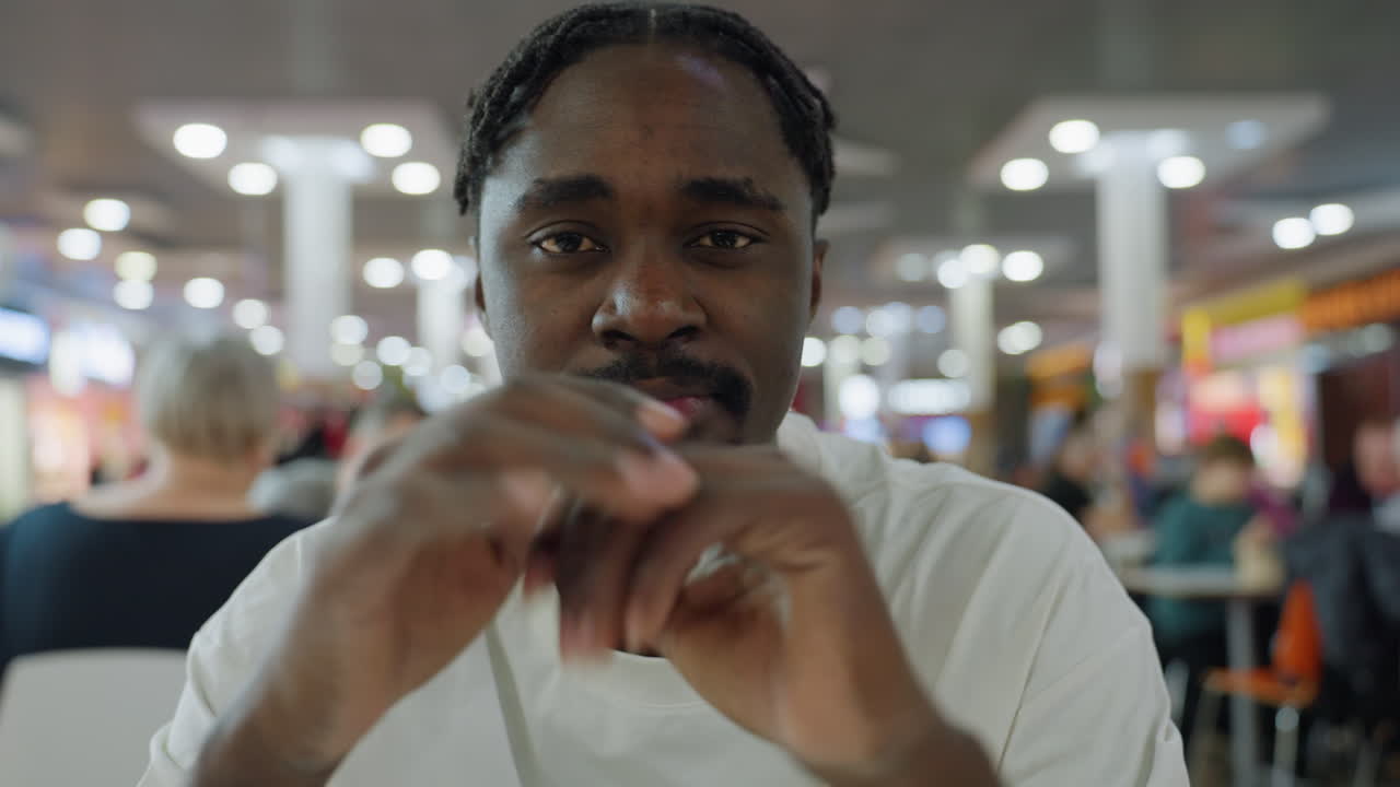 Young african man in white shirt sits indoors surrounded by people rubbing palms together in thoughtful mood while looking around, with bright lights and blurred background