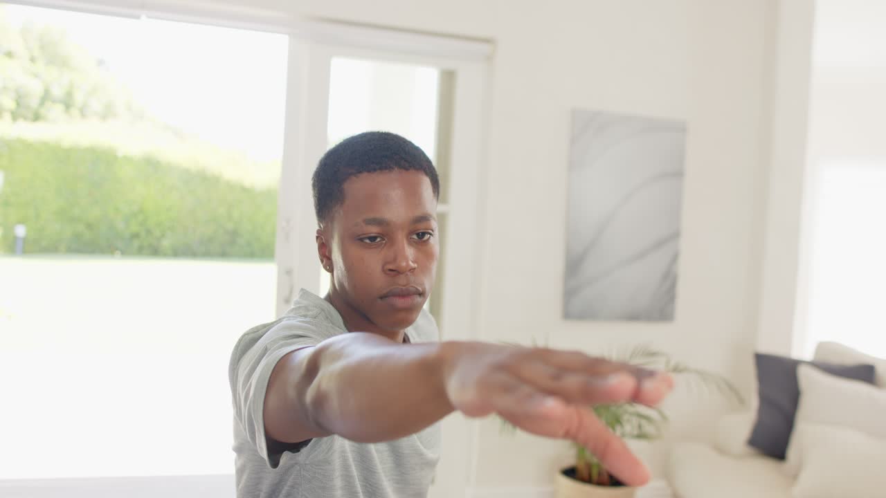 African American man following earbud instruction, adjusting extended arm slowly in living room