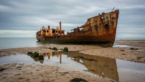 abandoned shipwreck on sandy shoreline reflecting in calm waters under a cloudy sky, captured in stunning detail with rust patterns and seaweed surroundings