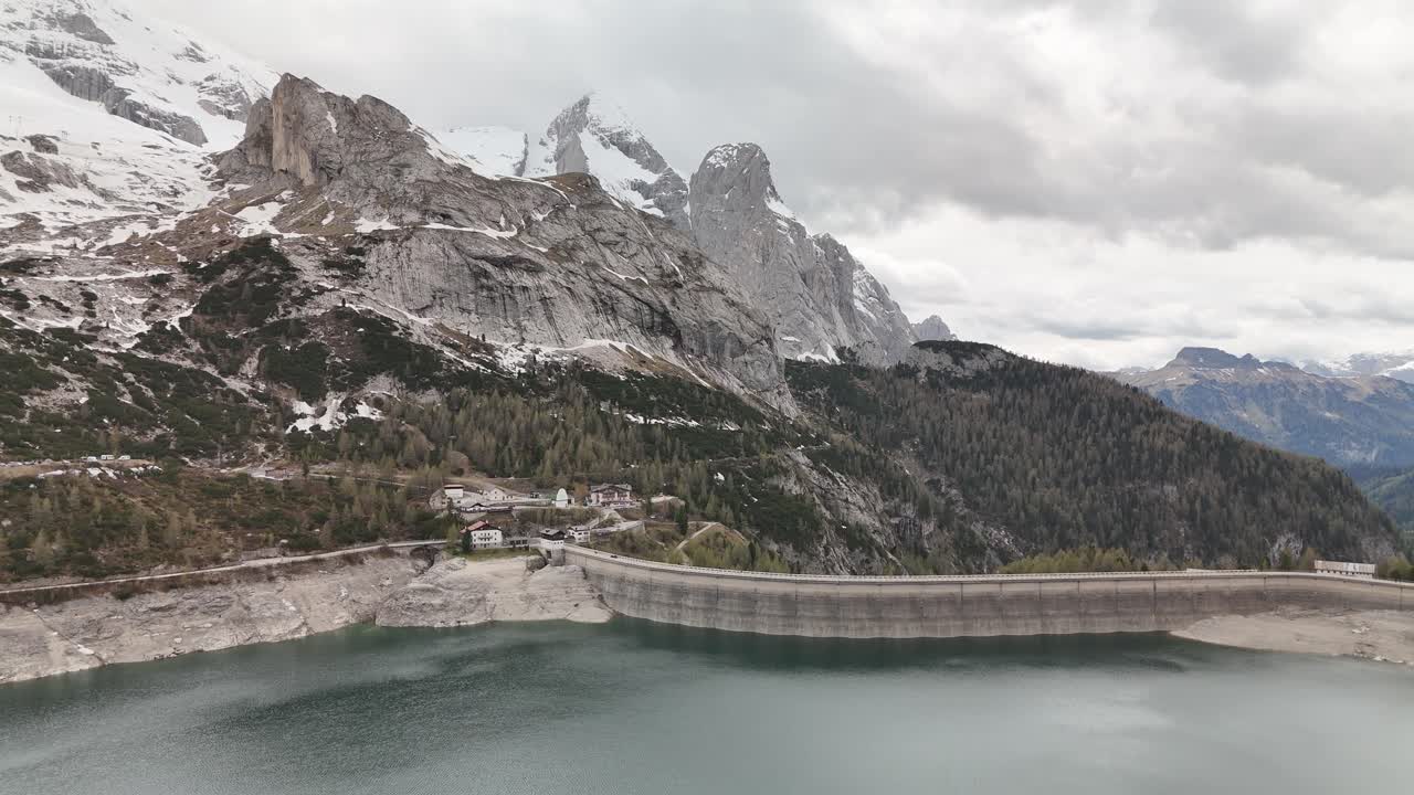 Aerial drone flight over Lago di Fedaia toward the dam and dramatic peaks of Marmolada in the Dolomites, Italy. Moody overcast light enhances the alpine textures and rugged mountain landscape