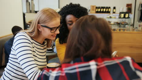 amigas afroamericanas y caucásicas hablando y viendo algo en una tableta sentadas en una mesa en un café