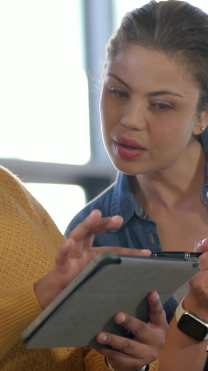 Vídeo vertical: Mulher de blusa mostarda rolando um tablet no escritório, enquanto uma pessoa vestida de jeans anota em um caderno.