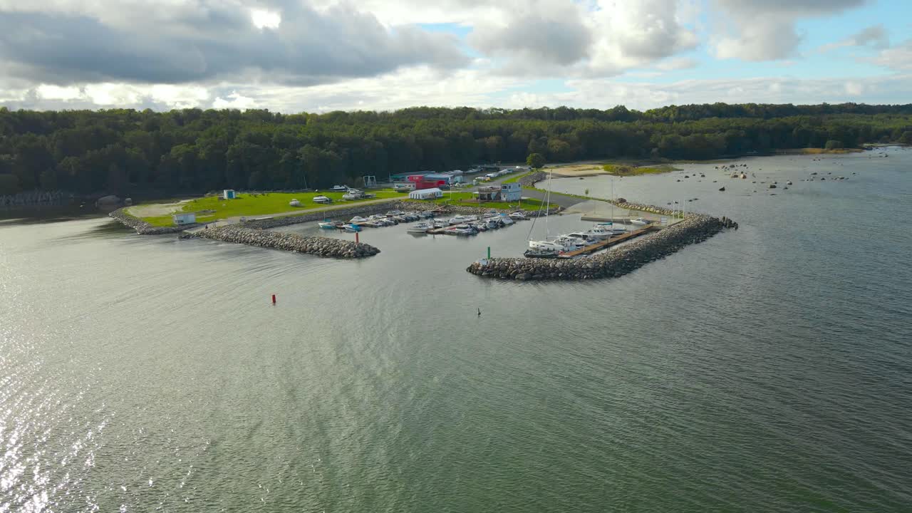 Aerial drone footage orbiting and flying around a luxury and small yach and sailboat harbor in countryside rural seaside shoreline where boats are docked on rocky piers during sunny summer day