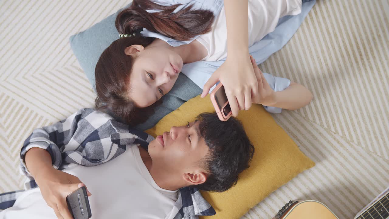 집에서 바닥에 카에 누워있는 동안 스마트폰을 사용하는 아시아 십대 커플의 가까운 상단 뷰. 전화 중독 커플이 함께 이야기하고 웃고 시간을 즐기고 있습니다.