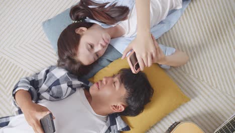 close up top view of asian teen couple playing smartphone while lying on carpet on the floor at home. phone addicted couple speaking, laughing, and enjoying time together