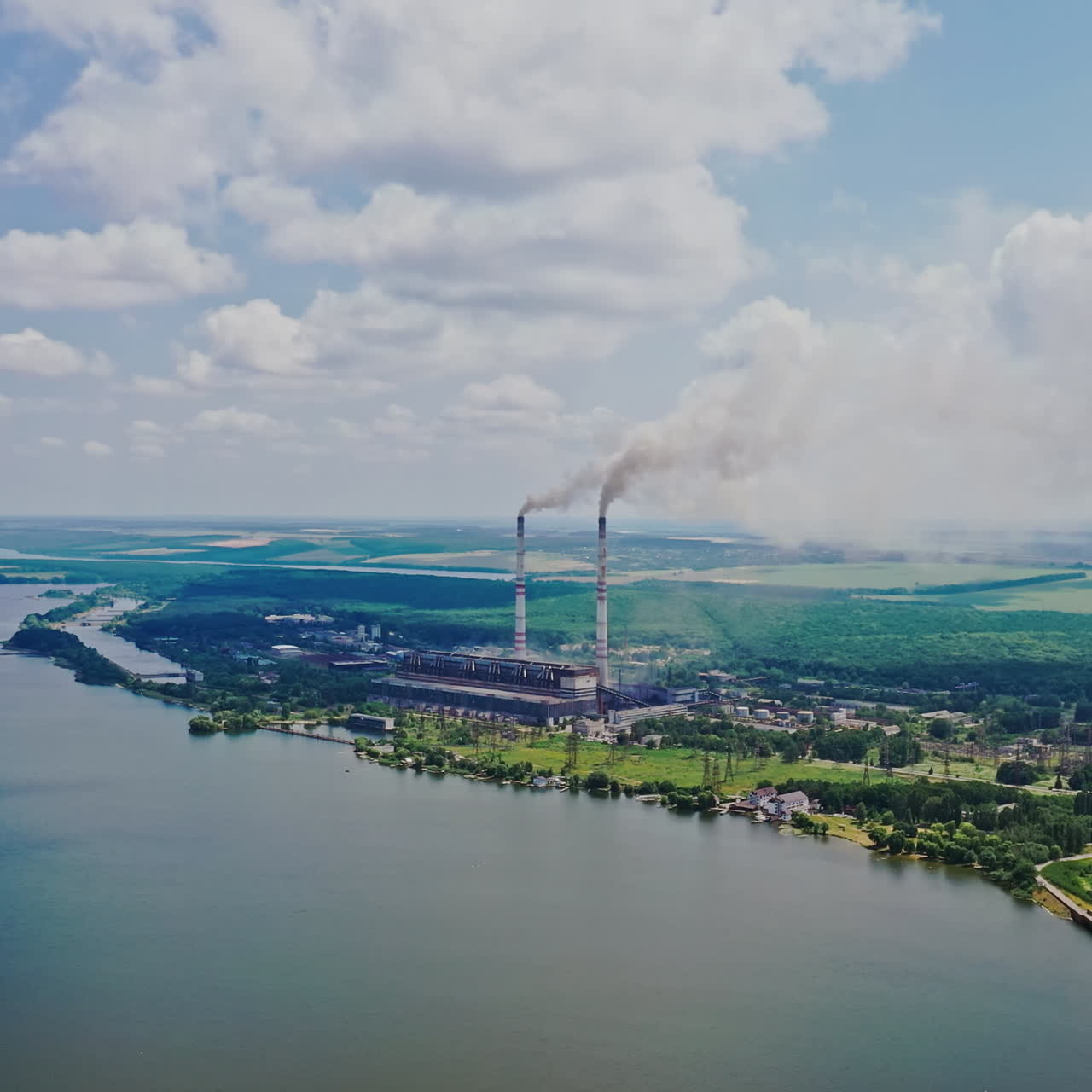 Aerial view of plant on the bank of the river in summer. View from above on natural background of beautiful river and factory with smoke from pipes. Slow motion. Camera moves up.