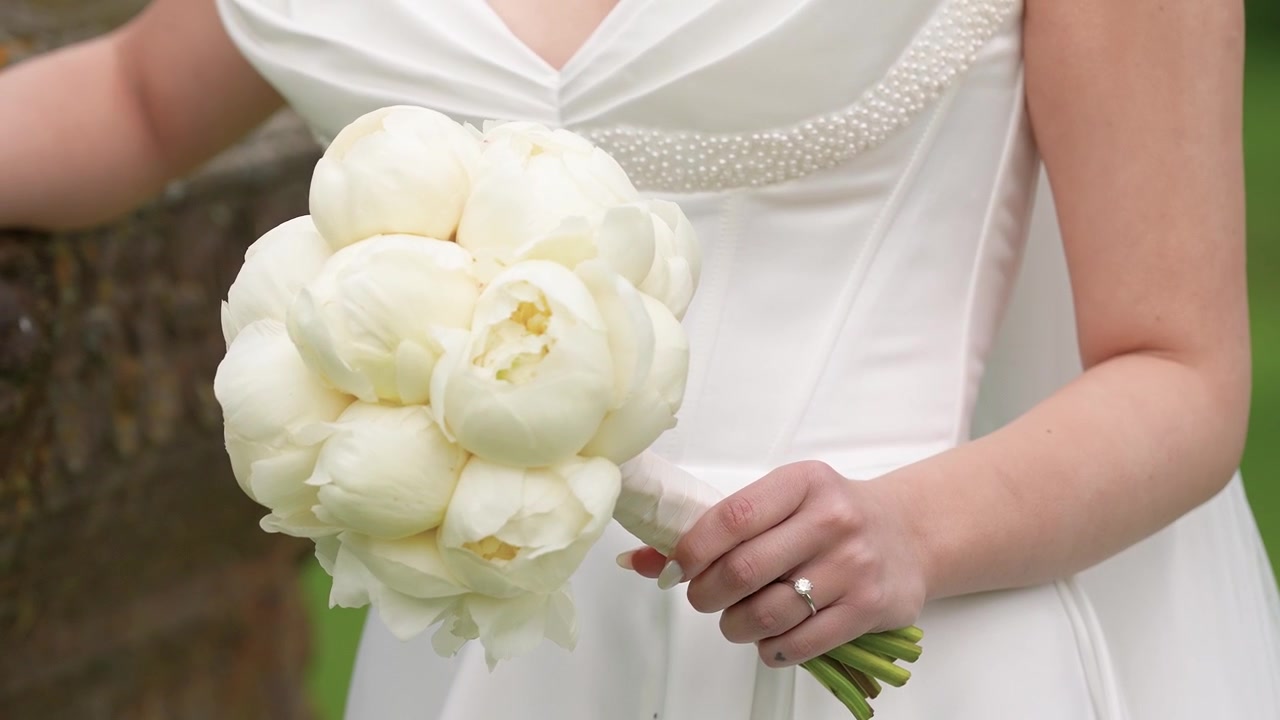 Elegante scatto ravvicinato di una sposa in abito bianco che tiene in mano un lussureggiante bouquet di peonie all'aperto, mettendo in mostra il suo anello di fidanzamento. La luce naturale del giorno e una profondità di campo ridotta esaltano la scena romantica.