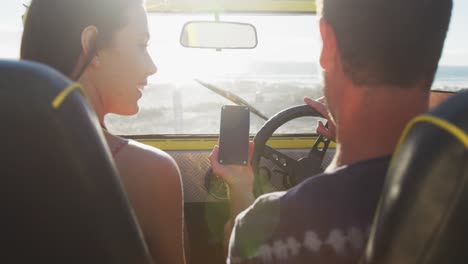 Feliz-Pareja-Caucásica-Sentada-En-Un-Buggy-De-Playa-Junto-Al-Mar-Usando-Un-Teléfono-Inteligente