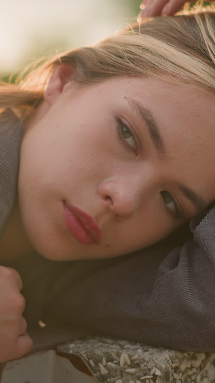 회색 옷을 입은 여자가 야외에서 팔에 머리를 고 머리카락을 부드럽게 만지고, 차분한 표정으로 생각에 빠져, 눈은 부드럽게 박이고, 배경은 약간 흐릿합니다.