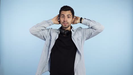 footage over blue background of a caucasian young man that keep combing his dark short hair and stares at the camera. feel unconfident and shy. guy has black headphones on his neck