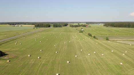 High-aerial-view-of-round-haybales-on-a-large-green-grassy-field-with-blue-skies-and-scattered-clouds-and-forest-in-the-background