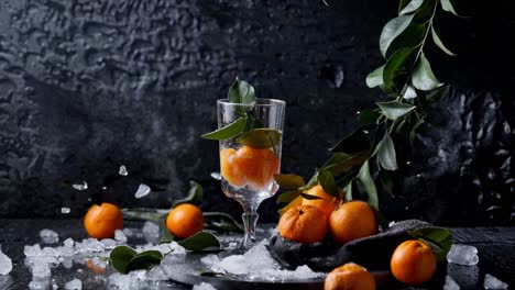 glass with fresh tangerines and ice along with a branch of tangerines and scattered ice on a dark background create a vibrant and refreshing still life