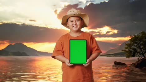 niño asiático con un sombrero sonriendo y mostrando una tableta de pantalla verde. niño investigador examina algo en un lago, concepto de aventura de turismo de viaje