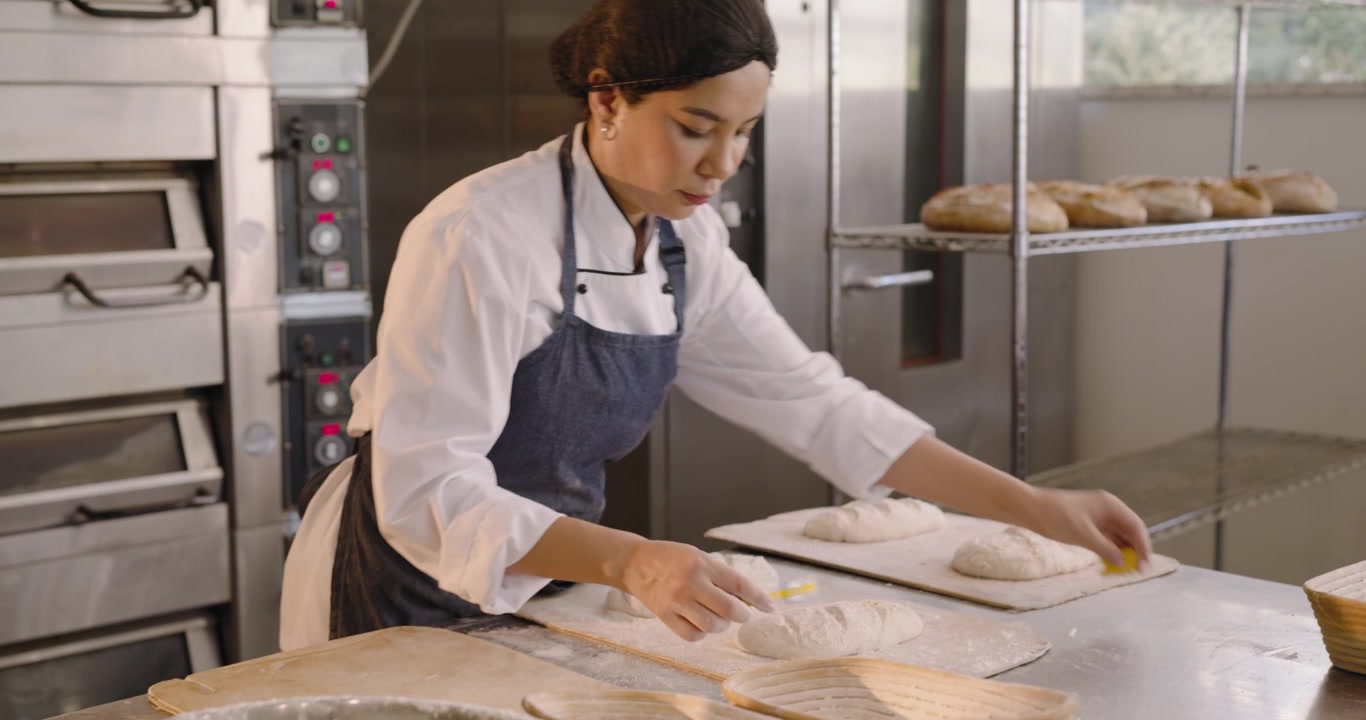 Padeira com uniforme de chef moldando massa descansada com uma lâmina de padeiro amarela em mesa de aço inoxidável, preparando o forno