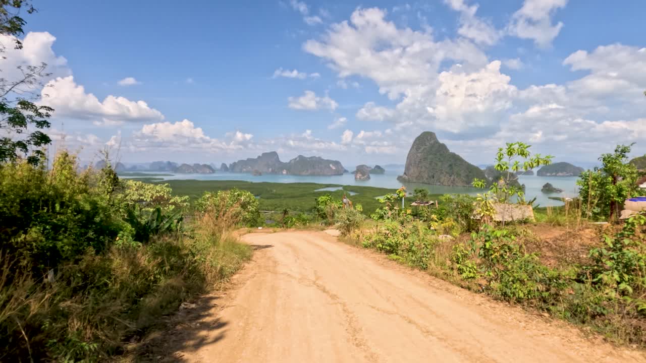 Camera moves along tropical dirt road revealing lush hills, dramatic limestone islands, and blue sky