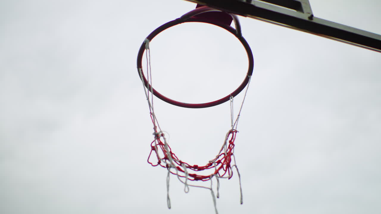 Canestro da basket consumato su cielo nuvoloso, rete lacerata che ondeggia su un campo vuoto evoca il ricordo di un giocatore di strada che si allena da solo, fischio dell'allenatore assente, bambino che sogna il tiro, sessione di allenamento al crepuscolo