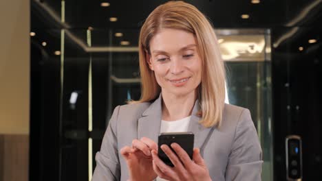 beautiful blonde businesswoman with mobile phone on the background of modern business center. pretty confident woman using smartphone, sending message, browsing internet, social media working indoor.