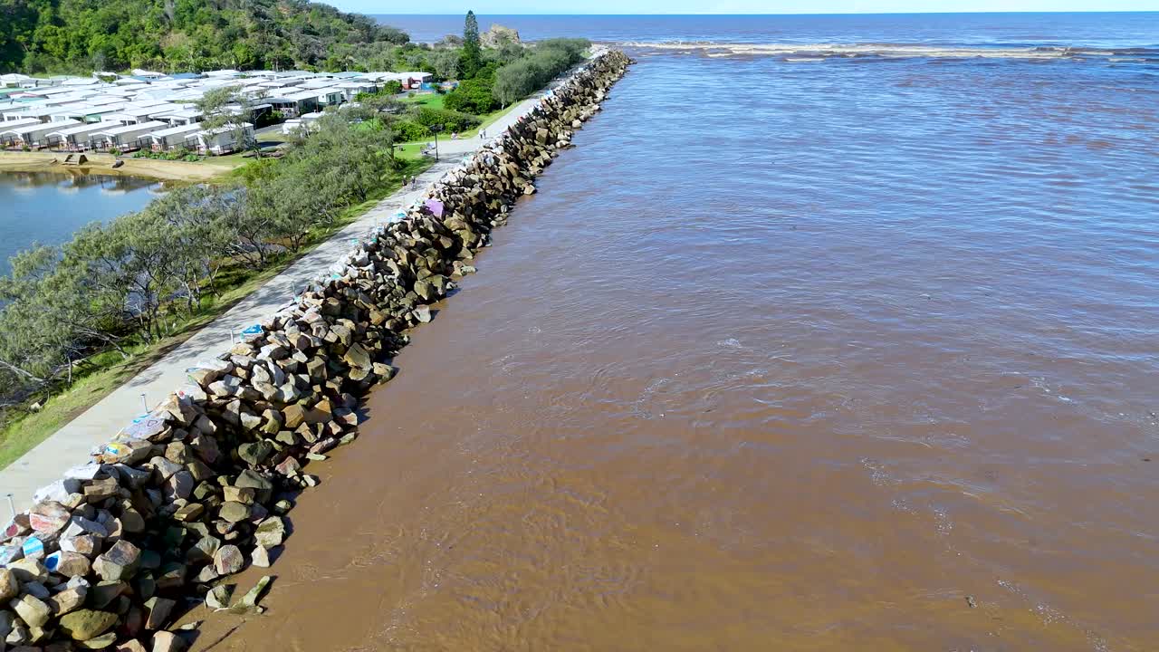 Aerial drone footage over stone breakwater separating brown river and green vegetation in daylight