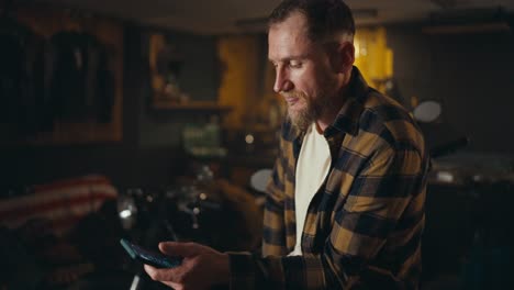 Confident-blond-guy-in-a-checkered-shirt-scrolls-social-networks-on-his-smartphone-in-a-workshop-garage