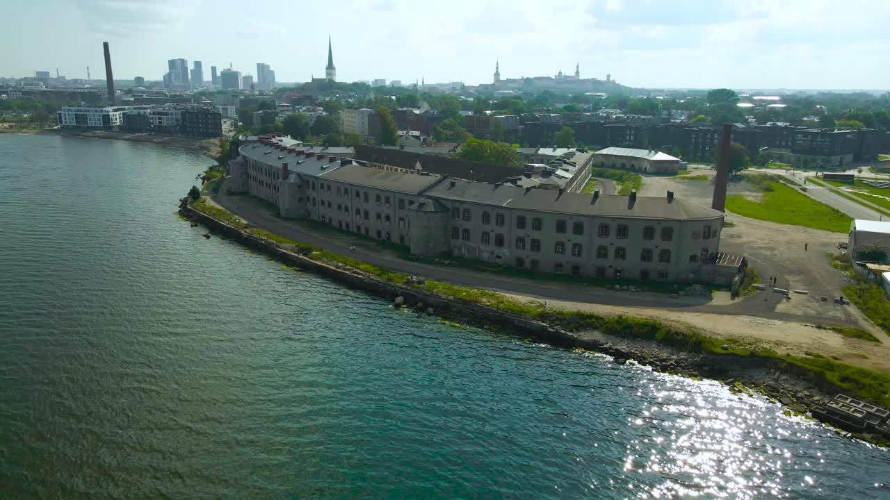 Aerial backward moving view glides beyond the city shore from Patarei sea fortress, old stone prison. Flying above gently rolling coastal waves along fort. Tallinn city skyline backdrop in summer day