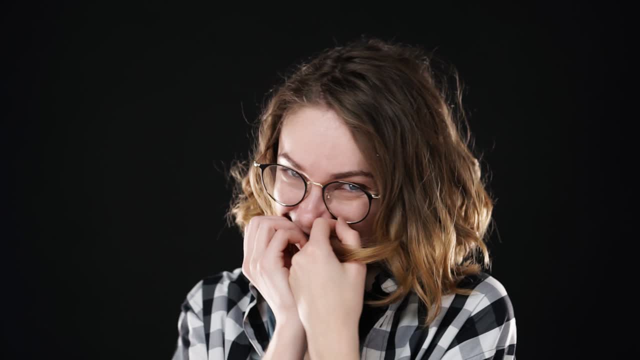 Close-up portrait of young european girl smiling humbly into camera over black background. Cheerfully smiling, embarrassed. Wearing eyeglasses and headphones on neck