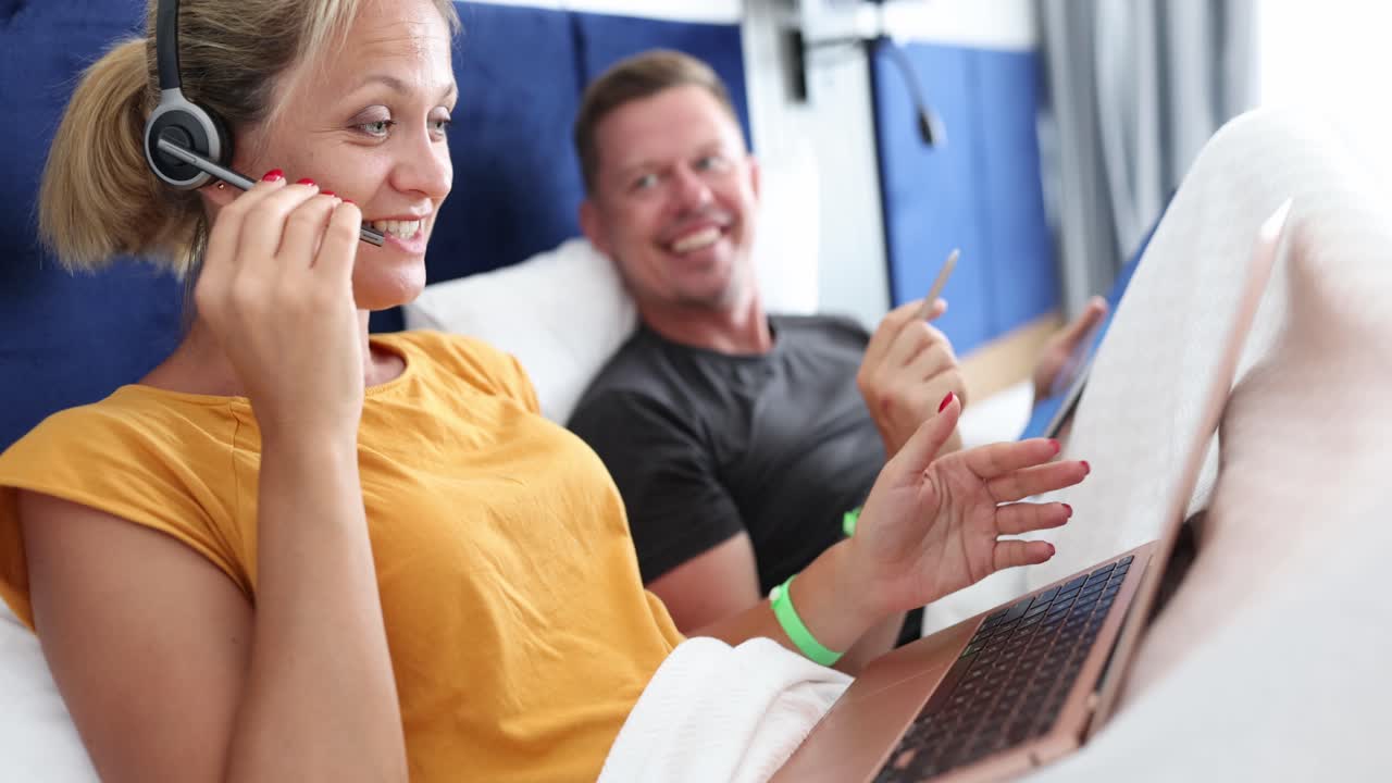 Woman on video call from bed with laptop and headset while man relaxes in background