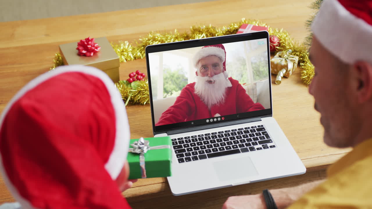 padre y hijo caucásicos con sombreros de santa usando una computadora portátil para una videollamada de navidad con santa en la pantalla