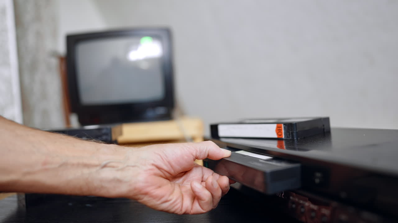VHS tape shoots out of the player. Senior man's hand takes a tape and puts on the desk. Close up
