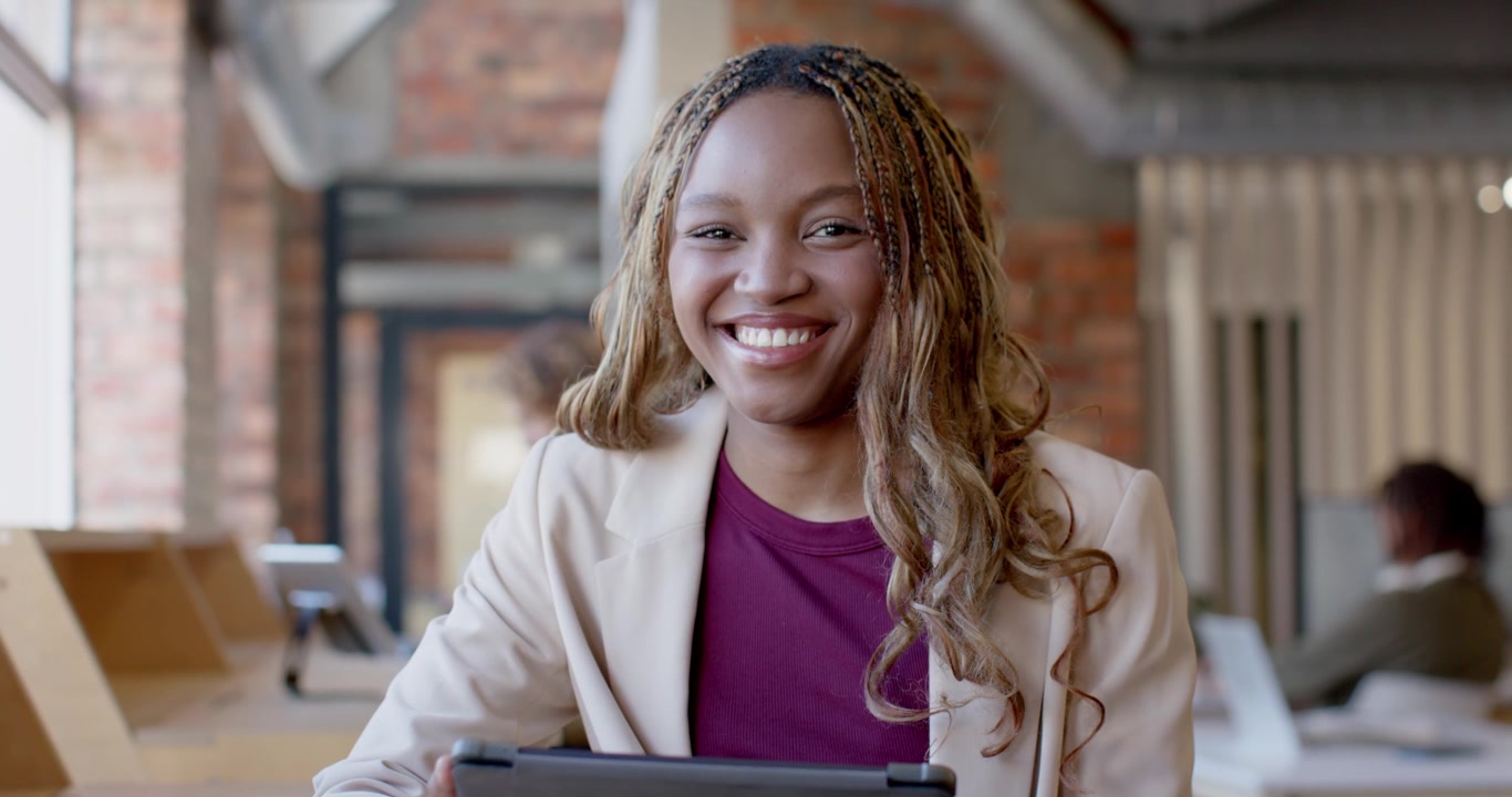 Donna afroamericana in blazer beige, che reagisce a un tablet e scoppia a ridere in ufficio.